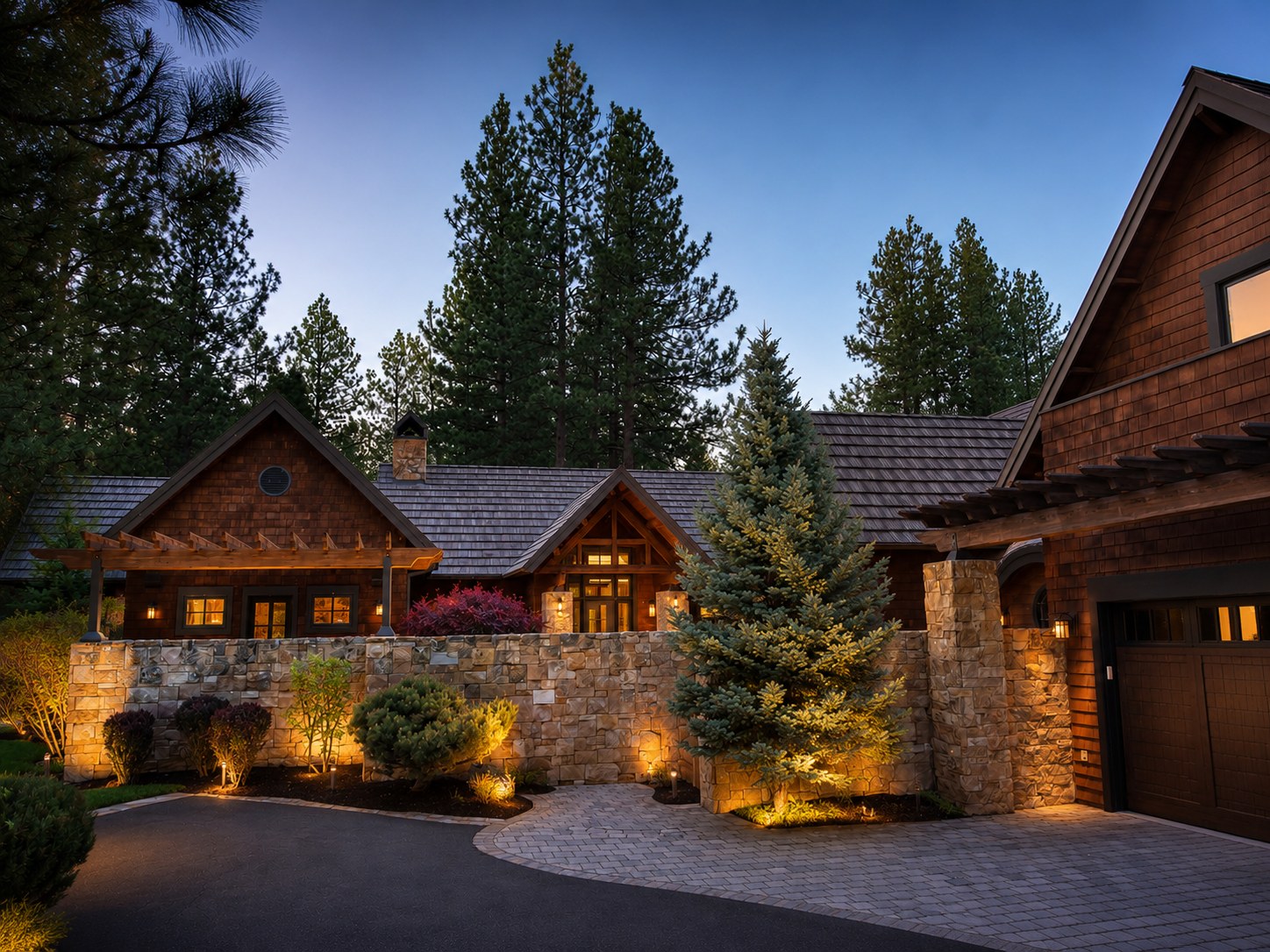 Twilight exterior of Broken Top Golf Course home — cedar shake siding, stone perimeter wall, illuminated landscaping, and ponderosa pine backdrop — LT Northwest Builders