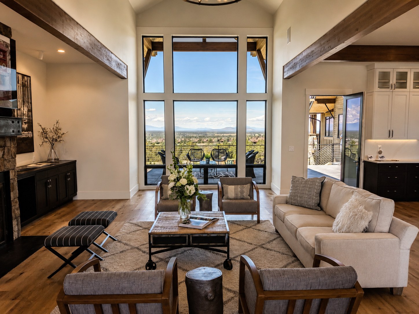 Custom home great room at Brasada Ranch Powell Butte Oregon built by LT Northwest Builders — vaulted cathedral ceiling with exposed dark wood beams, stone fireplace, hardwood floors, floor-to-ceiling black-framed windows with panoramic Cascade mountain and Central Oregon valley views, open to outdoor deck