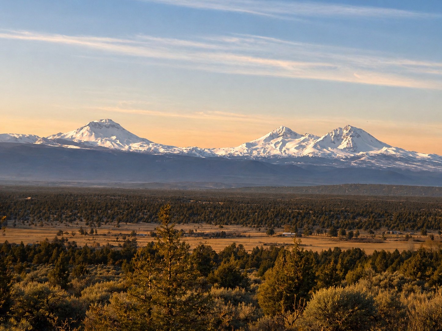 The Three Sisters mountains at golden hour in Central Oregon — snow-capped Cascade peaks rising above high desert sagebrush, juniper, and ponderosa pine landscape, photographed by Tom McClain of LT Northwest Builders